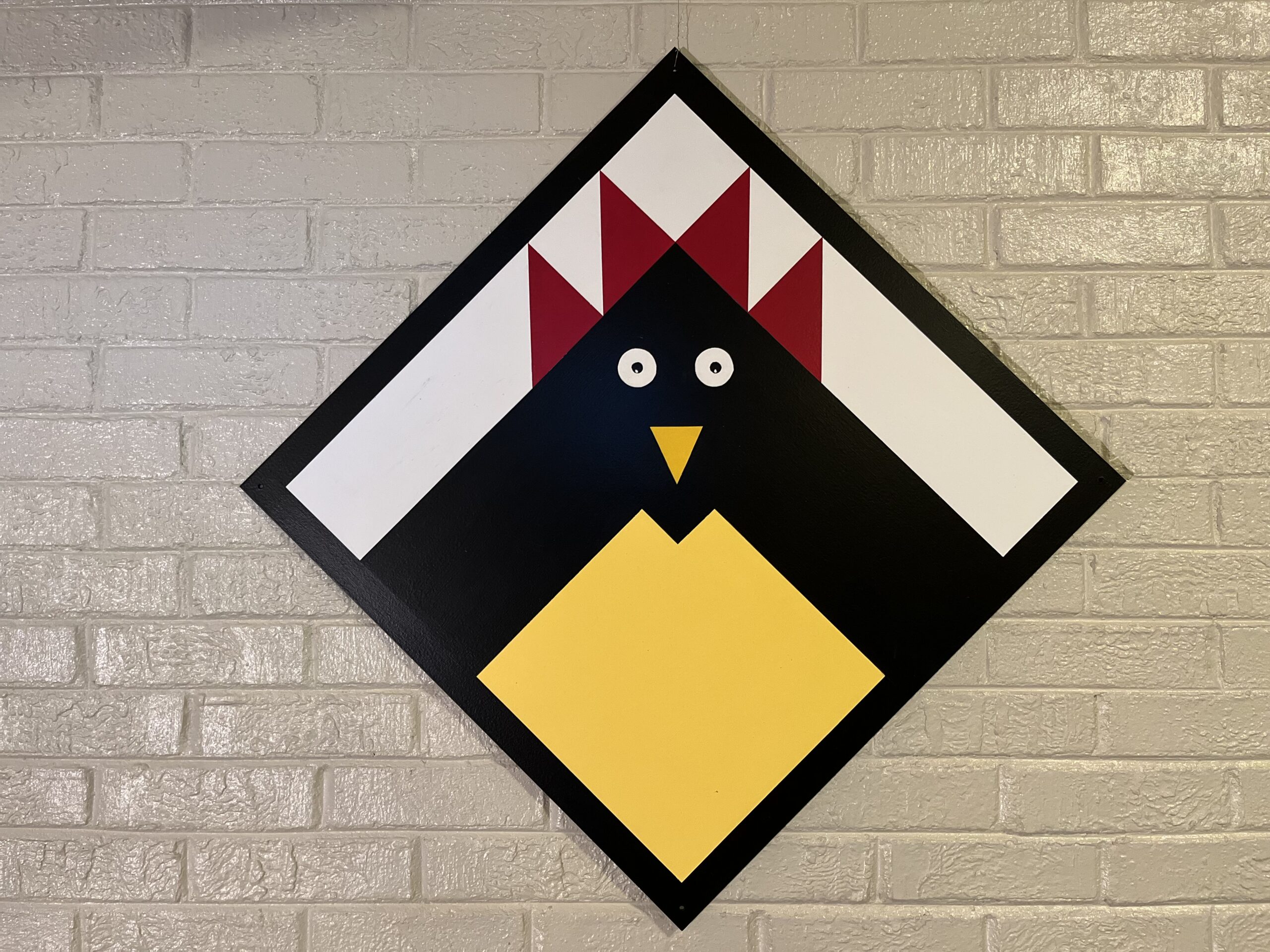 Colorful geometric art depicting a chicken, featuring a black body, yellow beak, and red crest, mounted on a light brick wall.