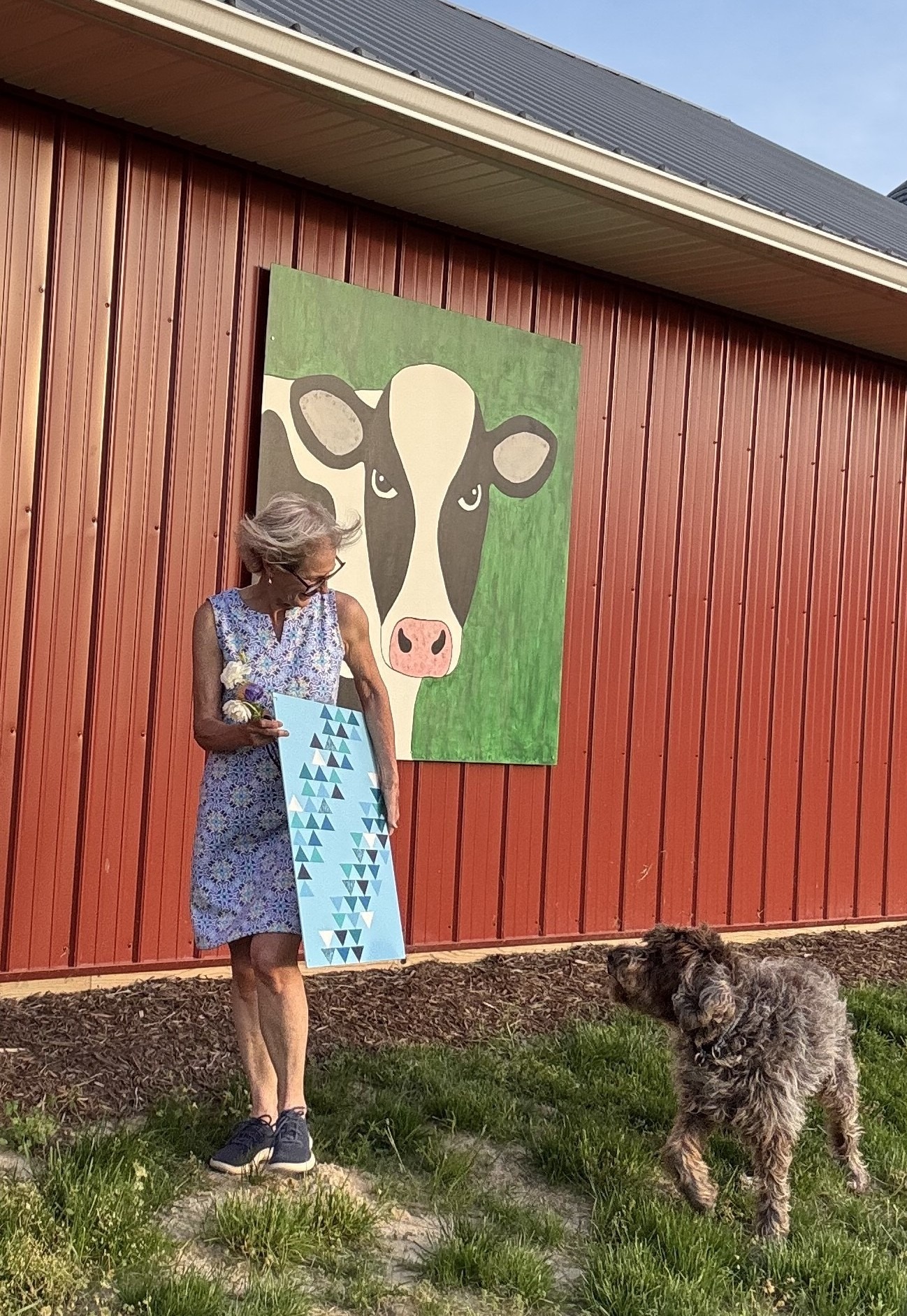 Woman in a floral dress holds a blue and triangle-patterned board beside a dog, with a large cow mural on a red barn in the background.